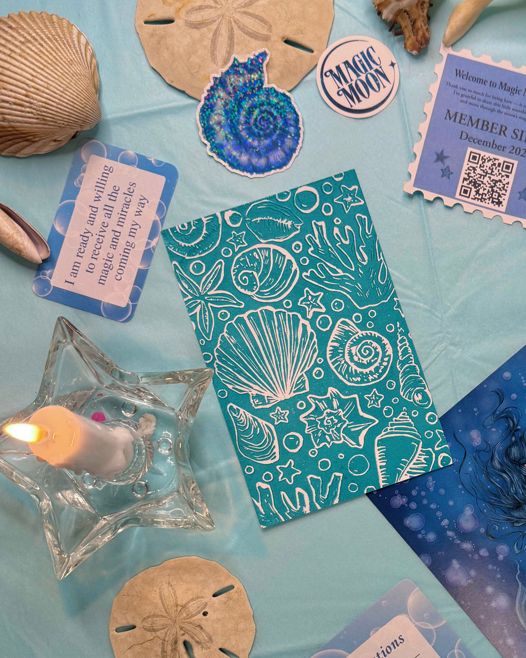 Teal linocut with shell designs surrounded by seashells, a candle, and other decorative items on a blue background.