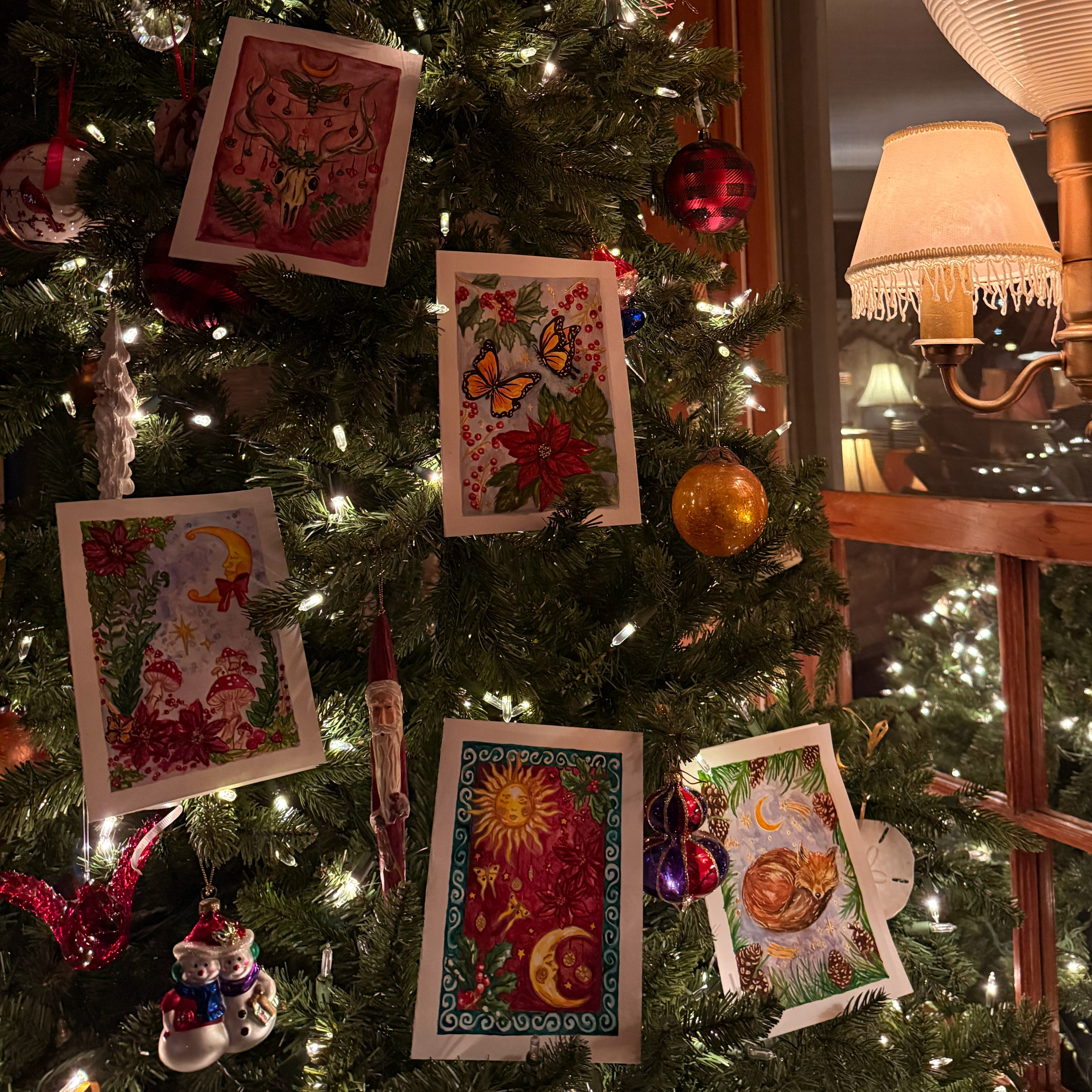 Decorated Christmas tree with watercolour christmas cards and ornaments, including a lamp and window in the background.
