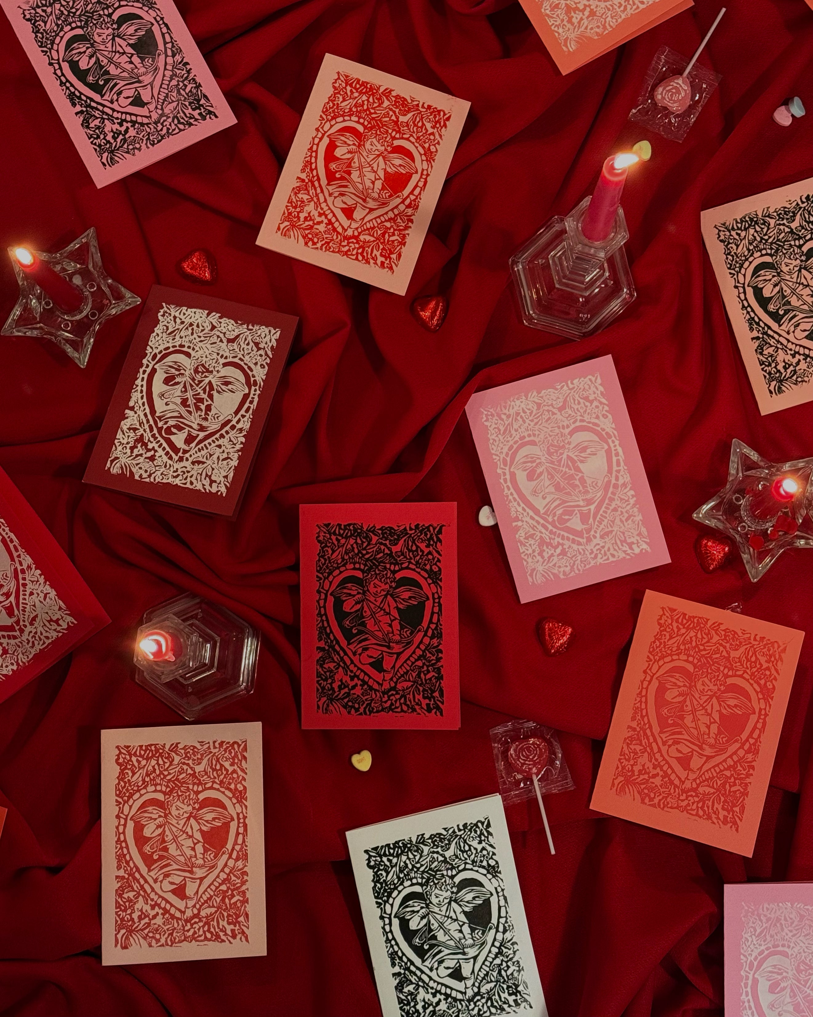 Decorative linocut cards with intricate cherub designs on a red fabric background with candles and stars.