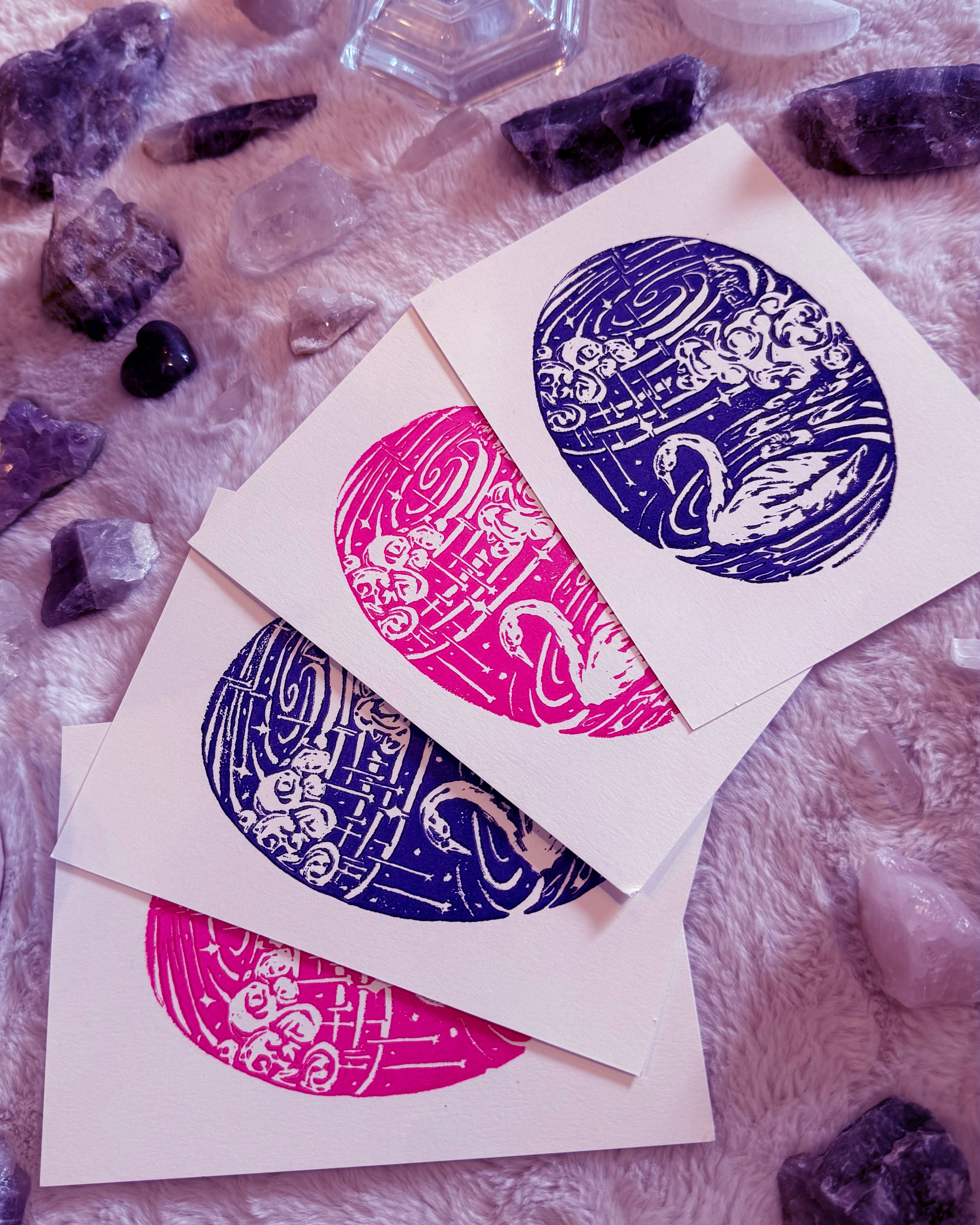Four circular linocut designs in purple and pink on white cards, surrounded by purple crystals.