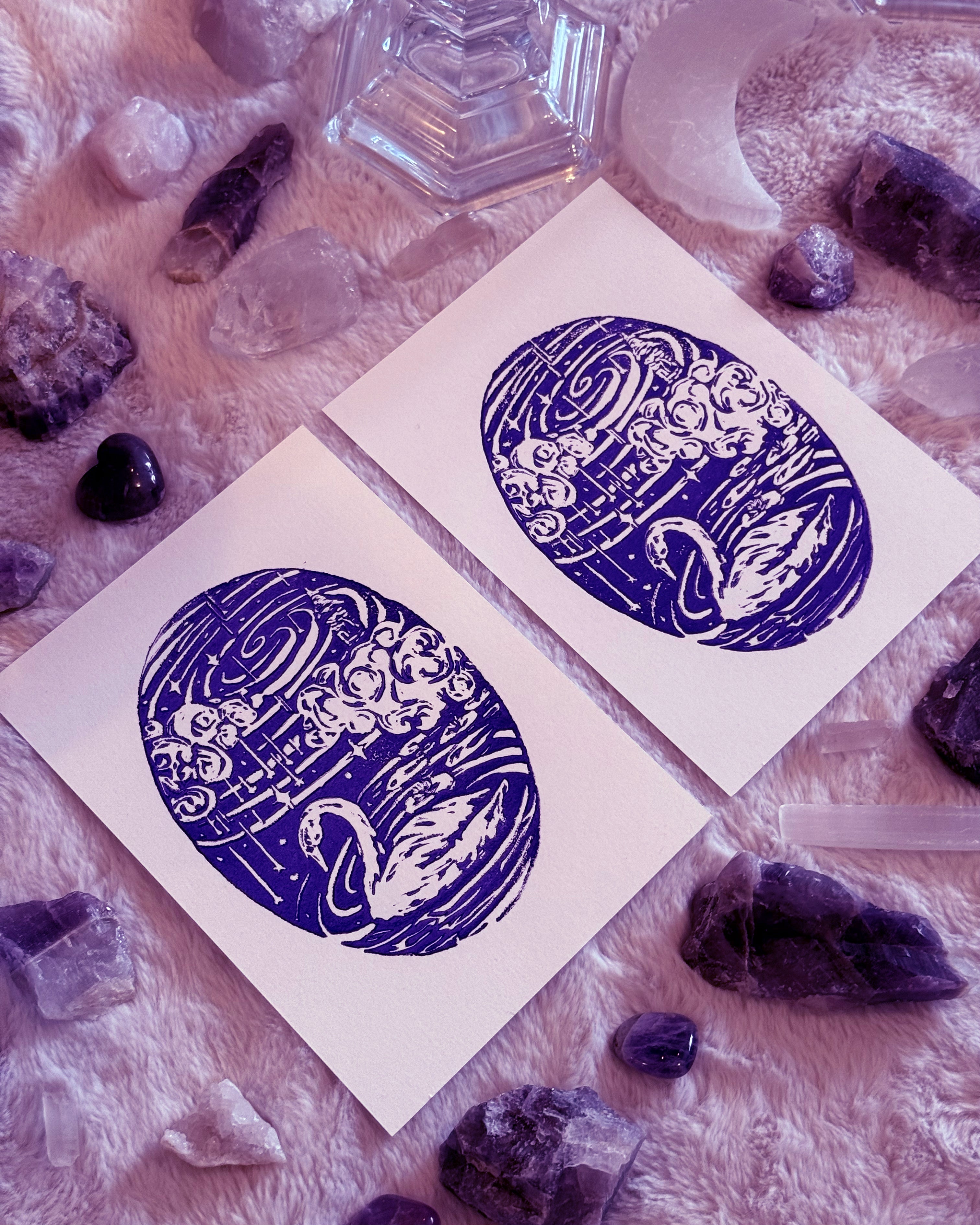 Two blue and white linocut circular swan designs on white paper surrounded by crystals and stones.