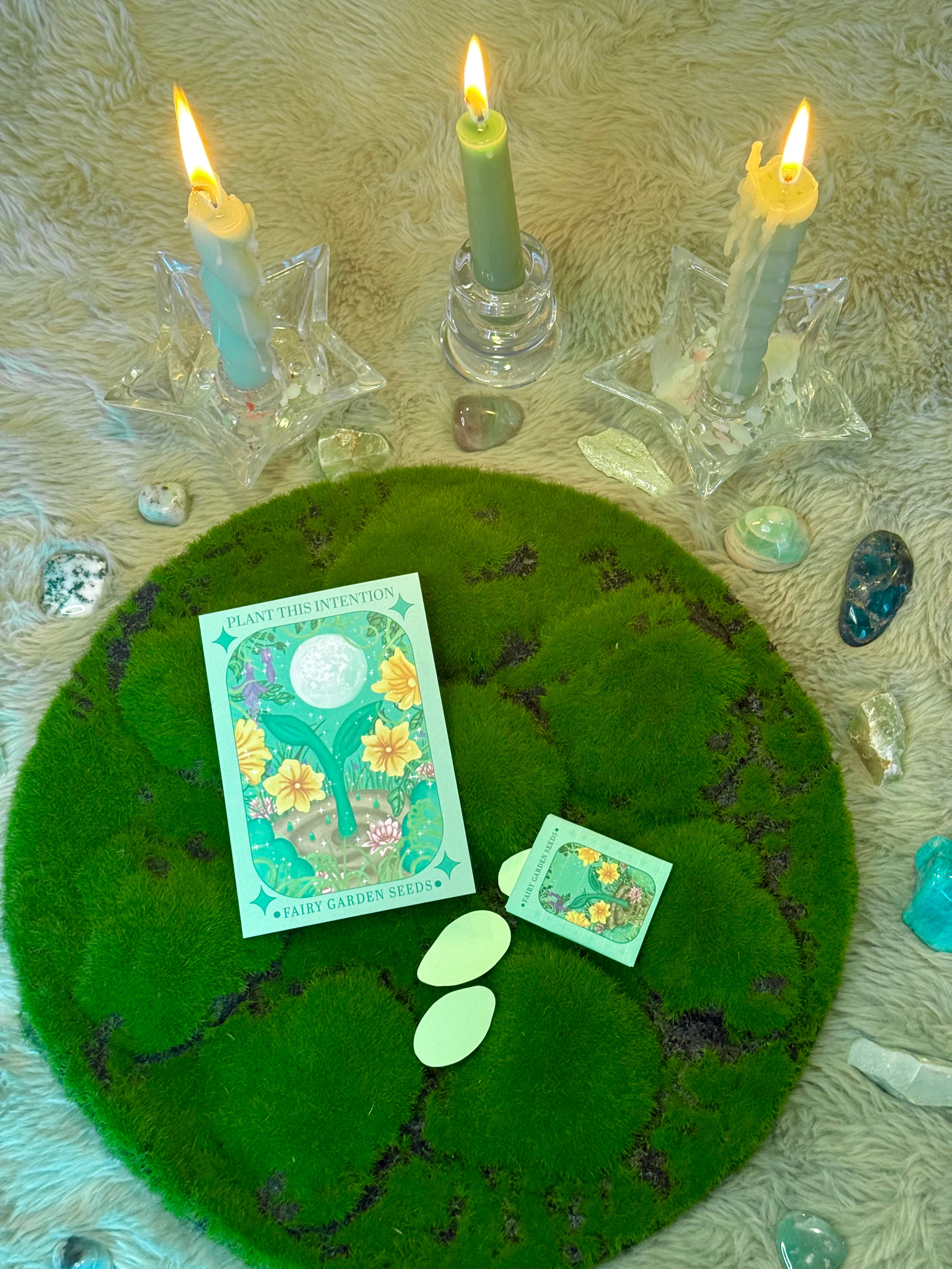 Three lit candles on a textured surface with a green card and small cards on a mossy circle.