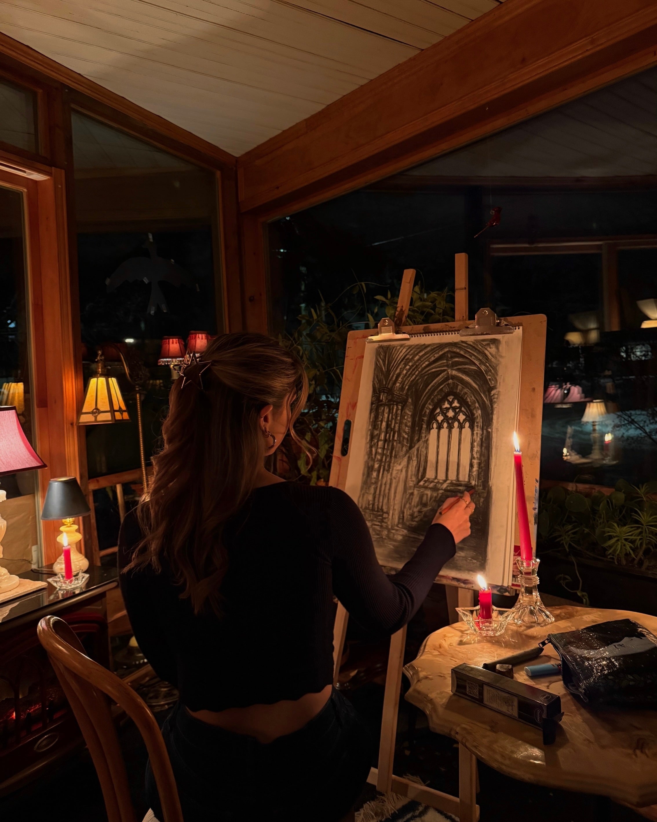 Person painting a cathedral scene on an easel in a dimly lit room with candles and lamps.